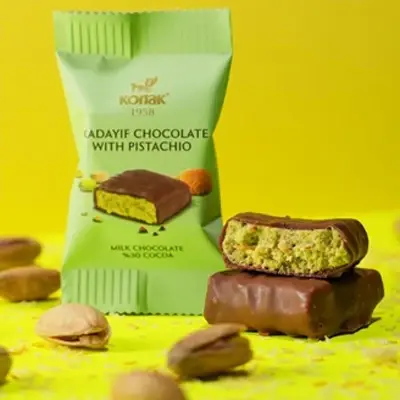 Unwrapped Konak Kadayif Chocolate with Pistachio bar displayed with packaging and scattered pistachios on a yellow background.