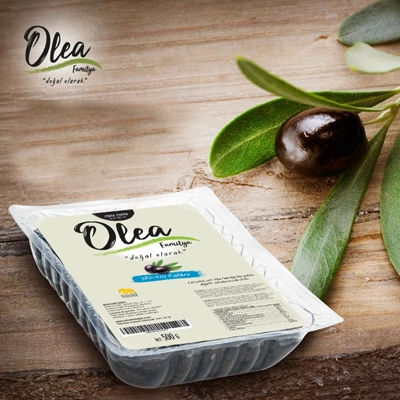 Olea black olive tray displayed on a wooden surface with a fresh olive and leaf beside it.